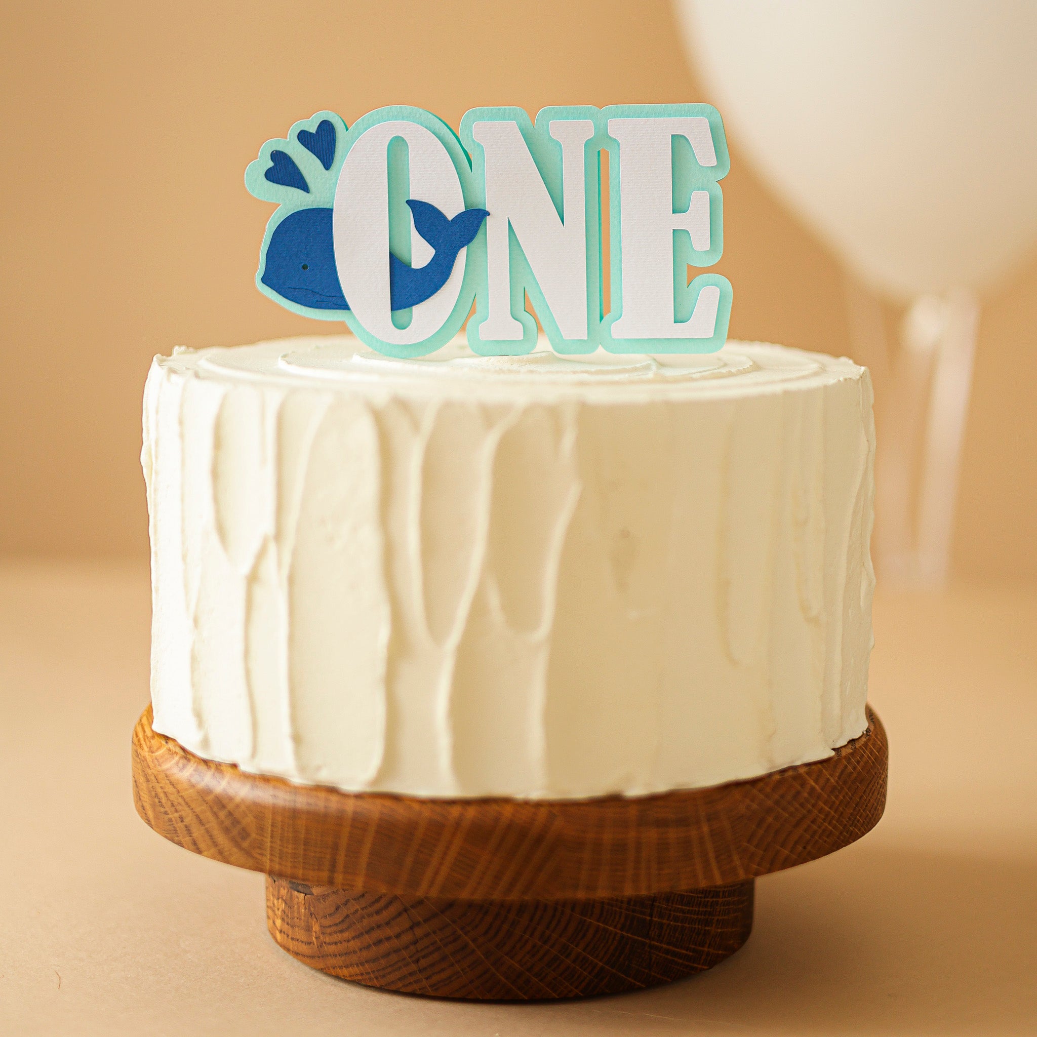 White cake with 'ONE' and whale decorations on a wooden stand against a beige background