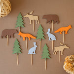 Eleven handcrafted woodland-themed cupcake toppers featuring cardstock silhouettes of orange foxes, brown bears, tan moose, grey rabbits, and green evergreen trees on wooden picks, arranged on a brown textured background.