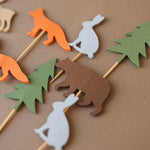 Eleven handcrafted woodland-themed cupcake toppers featuring cardstock silhouettes of orange foxes, brown bears, tan moose, grey rabbits, and green evergreen trees on wooden picks, arranged on a brown textured background.