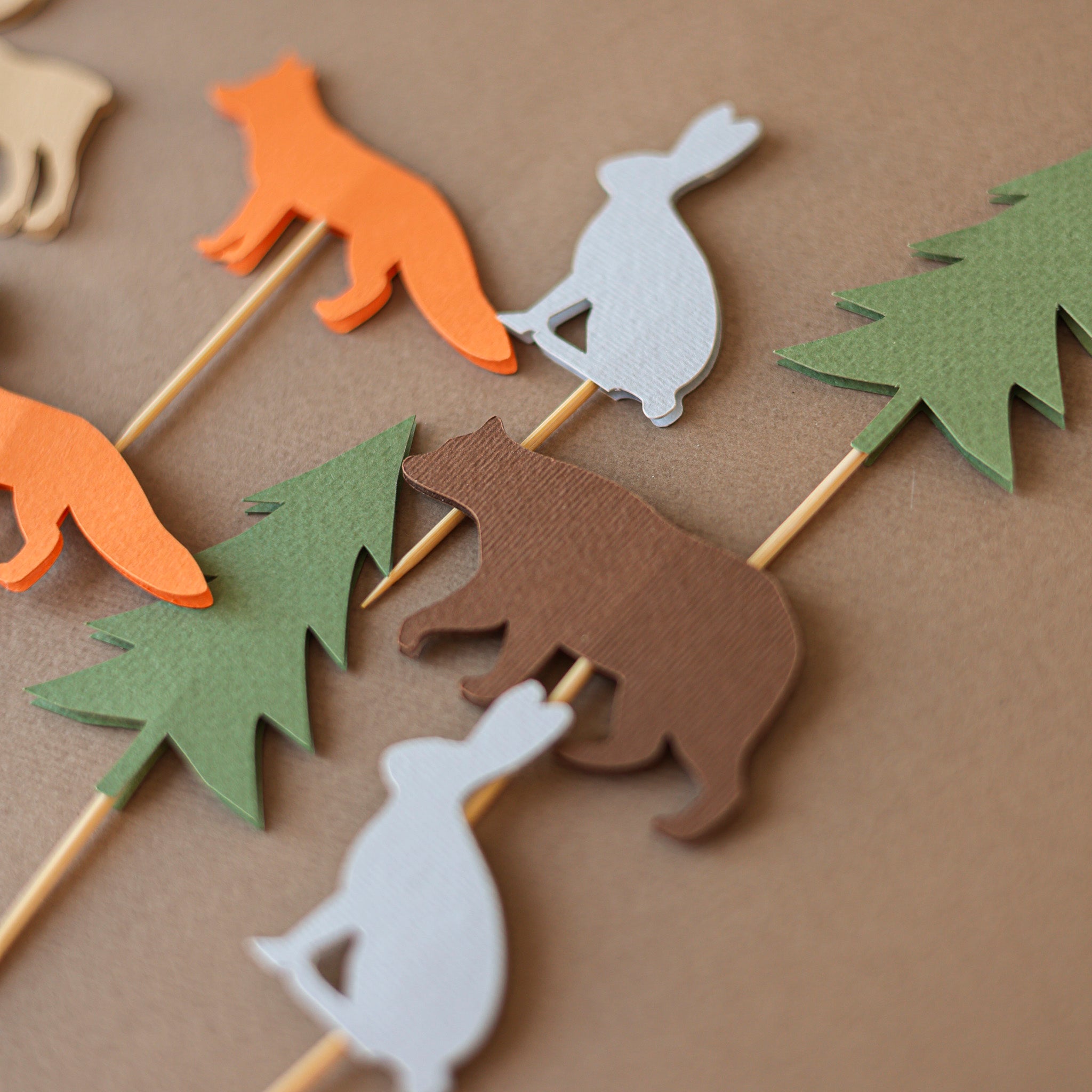Eleven handcrafted woodland-themed cupcake toppers featuring cardstock silhouettes of orange foxes, brown bears, tan moose, grey rabbits, and green evergreen trees on wooden picks, arranged on a brown textured background.
